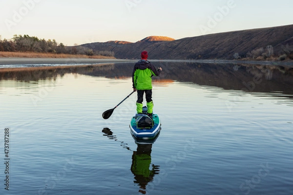 Fototapeta A man is rafting on the river on a supboard in the late autumn in the evening, looking into the distance at the mountains. SUP