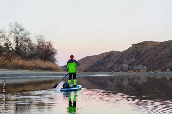 Fototapeta A man is rafting on the river on a supboard in the late autumn in the evening, looking into the distance at the mountains. SUP