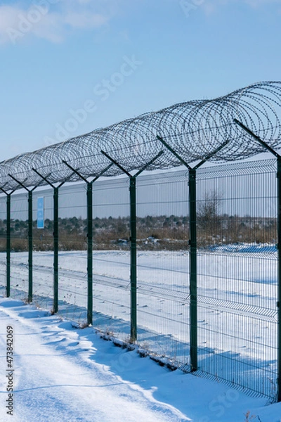 Fototapeta Iron fence with barbed wire in a snow-covered winter field against a blue sky. Fenced guarded territory, no access. Area protection and counteraction to illegal entry concept