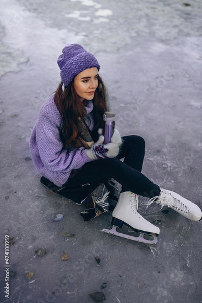 Fototapeta A beautiful woman is sitting on a skating rink and holding a thermos of hot tea in her hands. Toned in trendy Very Peri color of the Year 2022