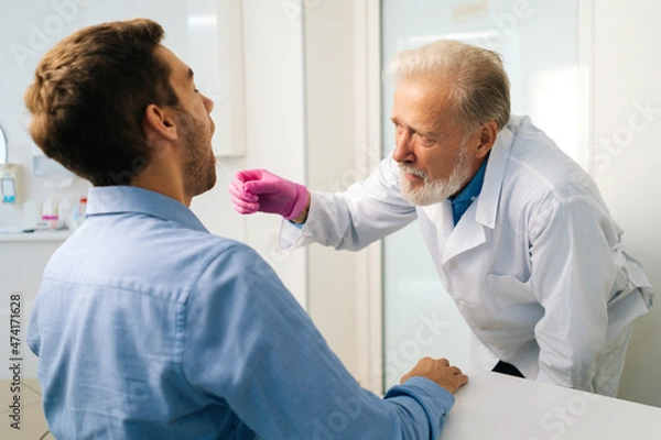Obraz Close-up view back to mature adult male doctor testing sick young man patient for possible coronavirus infection using throat swab in medical office. Man doing PCR test during corona virus epidemic.