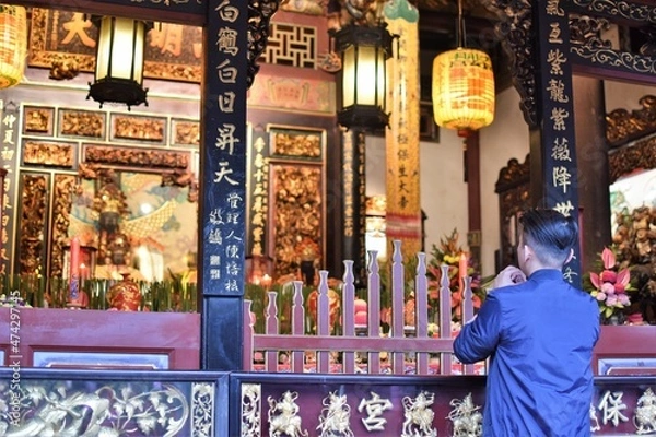 Fototapeta An Asian man prays at a shrine adorned with flowers and lanterns