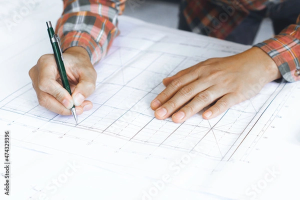 Fototapeta Young technician, engineer, designer holding a pencil drawing on a blueprint on his desk to create a project in the office..And free space to enter your text.