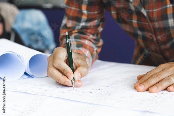 Fototapeta Young technician, engineer, designer holding a pencil drawing on a blueprint on his desk to create a project in the office..And free space to enter your text.