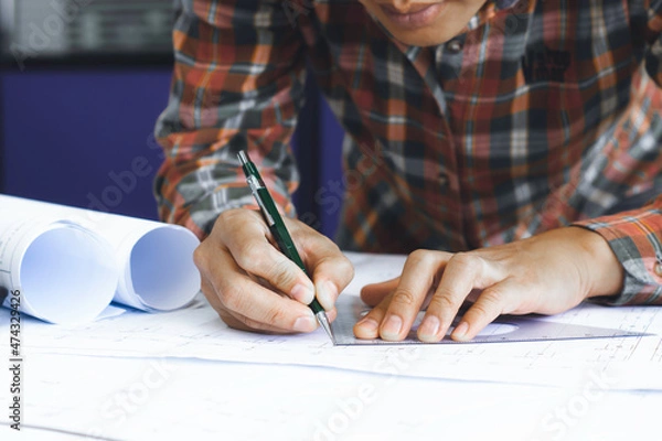 Fototapeta Young technician, engineer, designer holding a pencil drawing on a blueprint on his desk to create a project in the office..And free space to enter your text.