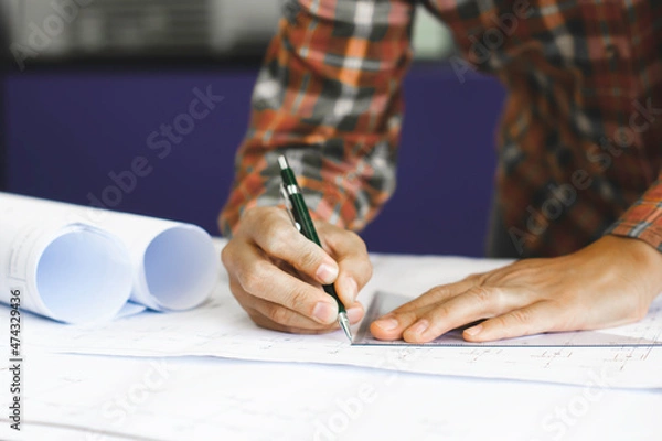 Fototapeta Young technician, engineer, designer holding a pencil drawing on a blueprint on his desk to create a project in the office..And free space to enter your text.