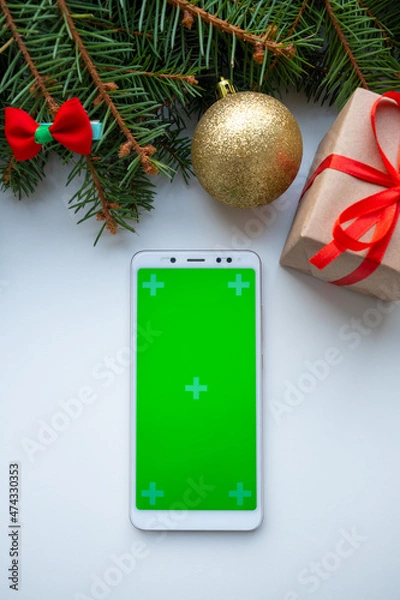 Fototapeta Smart phone mock up with Merry Christmas and Happy New Year decor on white background, green fir branches, balls and gift. Holiday decoration for app presentation. View from above, copy space.