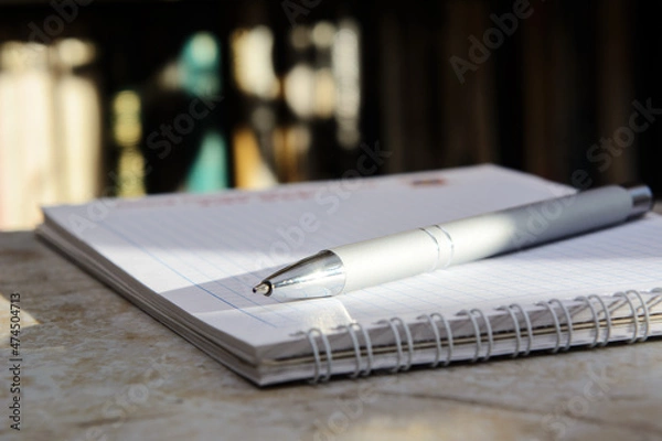 Fototapeta The ballpoint pen is located on the notebook. Background - books on the shelves. Solar lighting. Blurred colored background.