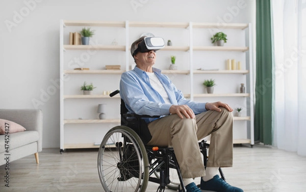 Fototapeta Happy senior man in wheelchair wearing VR glasses, playing online video game in cyberspace at home