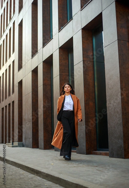 Fototapeta Asian business woman in long beige coat and hat walks down modern street on background of building in urban style
