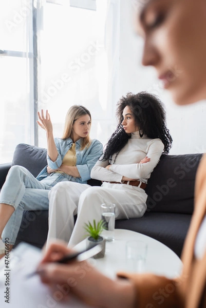 Fototapeta interracial same sex couple quarreling on sofa near blurred psychologist in consulting room