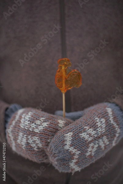 Fototapeta Caramel on a stick figure of a Cockerel in his hands in mittens.
