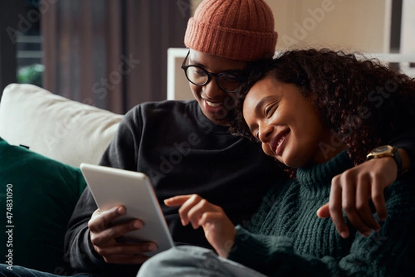Fototapeta Cute adult couple, multi-ethnic, smiling at tablet on sofa in modern apartment