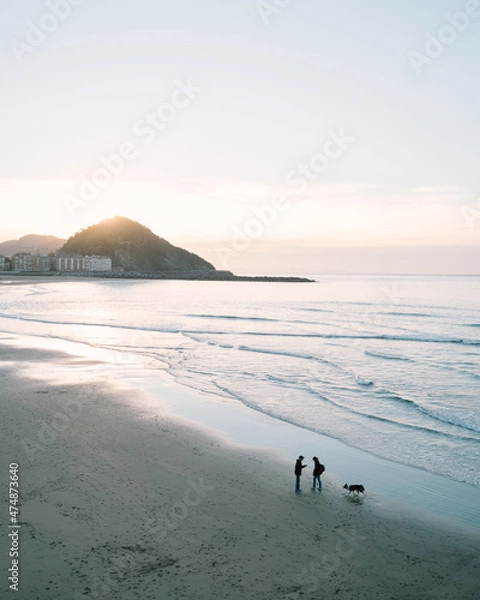 Fototapeta Couple talking on the shore of Zurriola Beach in San Sebastian during sunset while their dog is running around