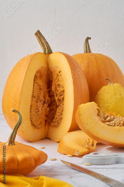 Fototapeta Several pumpkins on a light table, on a white background. One pumpkin is cut, pieces of pumpkin with seeds are next to it.