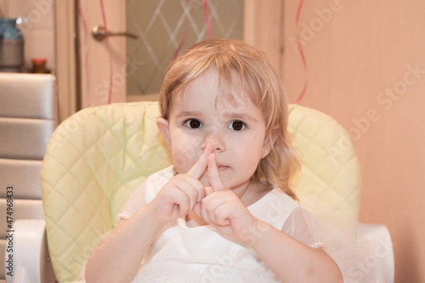 Fototapeta A little two-year-old girl sits on a highchair and shows that she is 2 years old