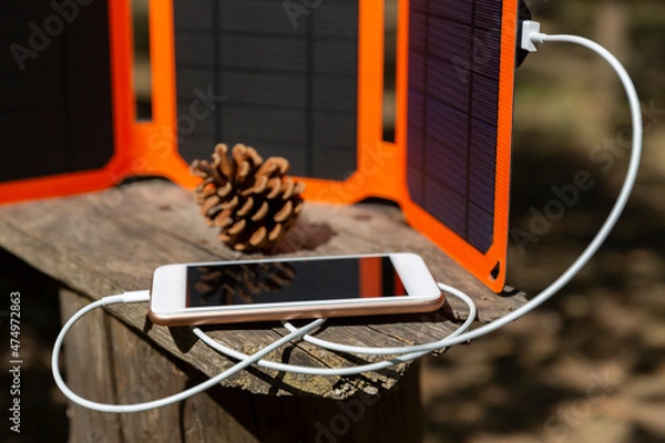 Fototapeta Smartphone is charging from solar battery in summer camp. Clean energy for using in camping. Gadgets are lying on the old wooden bench..