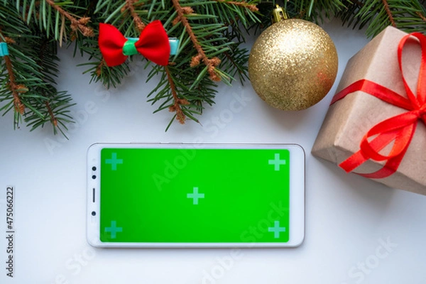 Fototapeta Smartphone mock up with Merry Christmas and Happy New Year gift on white background, green fir branches and balls. Holiday decoration for app presentation. View from above, copy space.