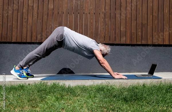 Obraz A sporty gray-haired man does yoga online using a computer, performs complex asanas