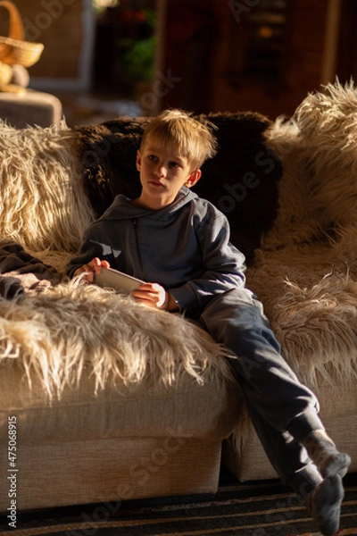 Obraz A blonde-haired child is sitting on a cozy sofa in a country house holding a smartphone in his hands