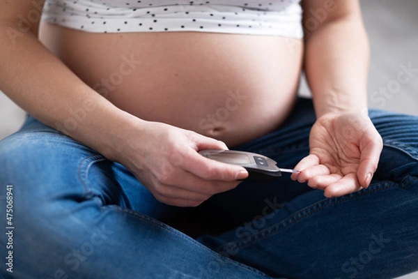 Obraz A pregnant woman measures her blood sugar level by puncturing her finger with a lancet while sitting on the floor