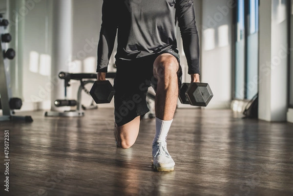 Fototapeta Young athlete holding dumbbells and exercising in the gym. He exercises to build muscle strength.