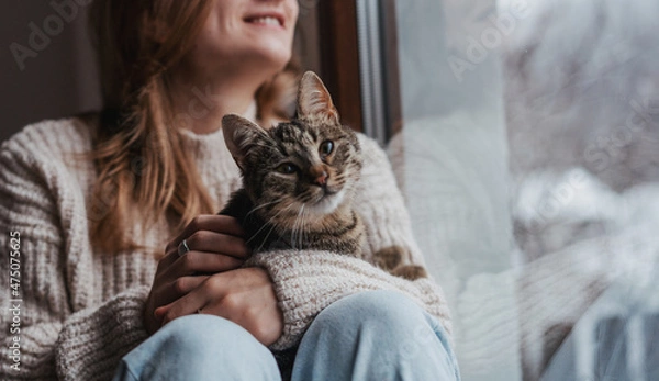 Fototapeta Young cheerful girl sitting at home on the windowsill in a warm sweater playing with a gray cat on a winter day