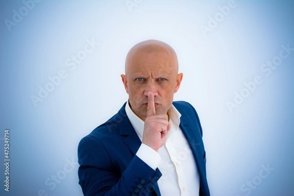 Obraz A brutal middle-aged man in a business suit asks for silence. A bald man with a shaved head presses his finger to his lips and demands to close his mouth. Concept: silence.
