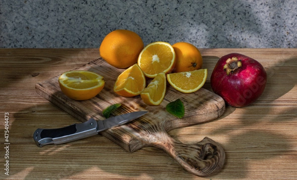 Fototapeta Still life: a group of fresh oranges, whole and cut, lies on an old figured wooden board, and next to it is a ripe pomegranate and a knife.  Drawing with light and shadow. Selective focus