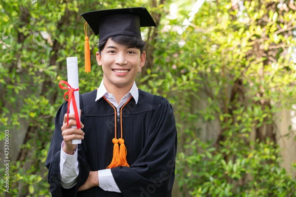 Fototapeta In the graden, a happy young Asian man wears graduation gowns and has a diploma.