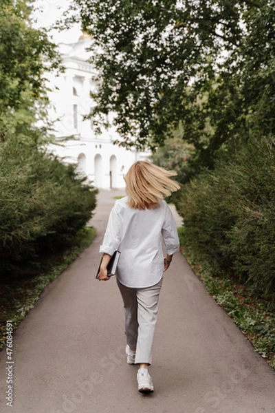 Fototapeta blonde in white clothes walks through the park to the university with a book in her hands green plants trees white building. vertical