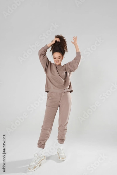 Obraz Beautiful slim model girl in a sports outfit and trendy sneakers pulling up her hair and posing on the white background