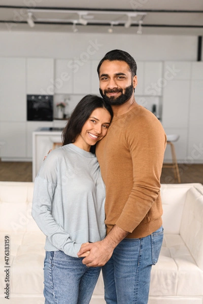 Obraz Smiling man hugging his charming woman. Two people standing and joyfully looking at the camera. Young Indian couple happily spending time in cozy modern kitchen at home