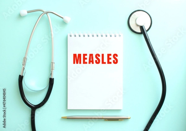 Fototapeta On a blue background, a stethoscope and a white notepad with the words MEASLES.