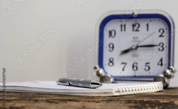Obraz There is a ballpoint pen on a notebook lying on an old blackboard. There is an alarm clock in the background. Concept business
