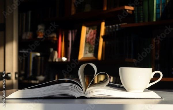 Fototapeta Background - bookshelves lit by the sun. An open book is on the table. The pages are folded in the shape of a heart. On the right is a cup of coffee.