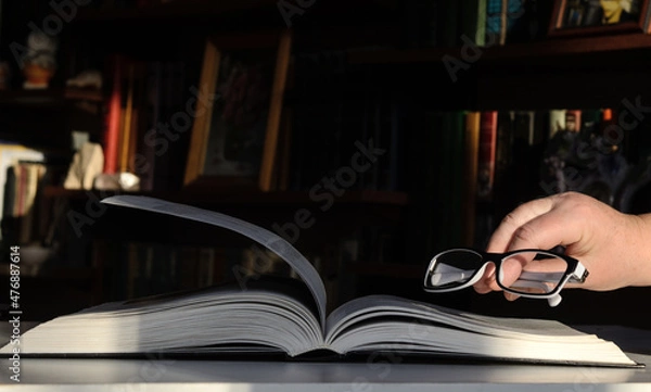 Fototapeta Sunlit bookshelves in the background. An open book is on the table. Glasses in the hand of the woman, whom she puts on the books.