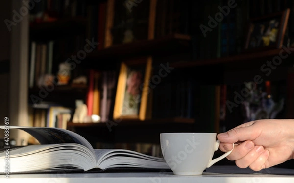 Fototapeta Background - bookshelves lit by the sun. An open book is on the table. Woman's hand with a cup of coffee