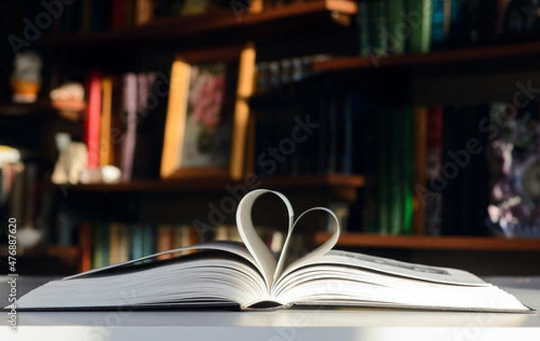 Fototapeta Background - bookshelves lit by the sun. An open book is on the table. Pages are folded in the shape of a heart.  Concept - Valentine's Day