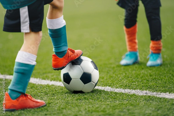 Fototapeta Close up image on soccer player legs with classic ball. Football background of soccer field and players in a game. Boy in sports shoes kicking ball.