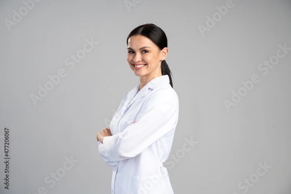 Fototapeta Side view of the smiling female doctor in lab coat with arms crossed looking away and posing against grey background. Medicine concept