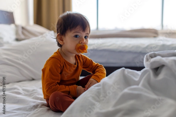 Fototapeta A one-year-old child with a pacifier in his mouth is sitting on the bed in the bedroom.