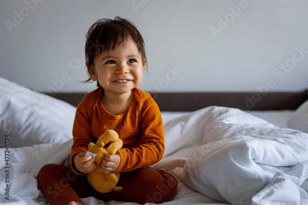 Fototapeta Portrait of a happy little child with a teddy bear sitting on the bed.