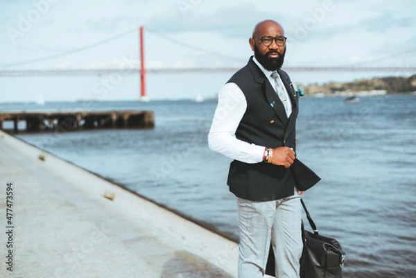 Fototapeta A businessman holding a messenger bag near a river, a red suspension bridge in a blurred background. A bald bearded black guy in a suit standing on the pier looking away, a copy space area on the left