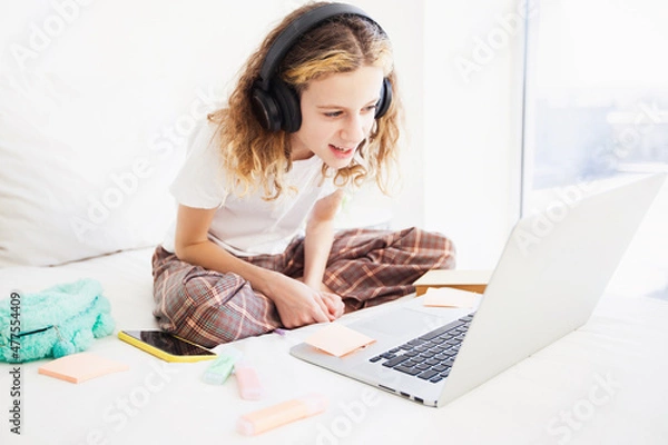 Fototapeta a girl child on the bed is looking at a laptop. The child is wearing headphones and waving at someone on the screen