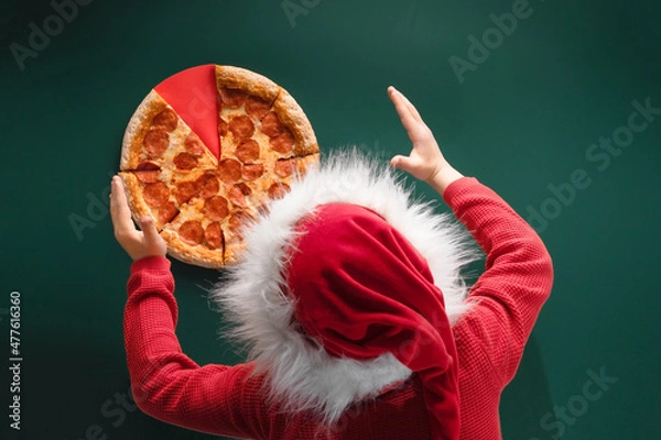 Obraz Top view of Pepperoni pizza and back of boy in red santa claus hat with fluffy fur  and on green background. The concept of a holiday, a Christmas Time to eat
