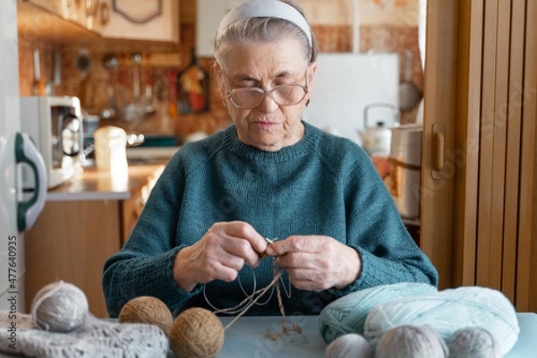 Fototapeta An elderly lady of 80 years old is engaged in an interesting hobby, knitting a brown jacket with knitting needles. Hobbies that you can do at home. A blurry image of the kitchen.