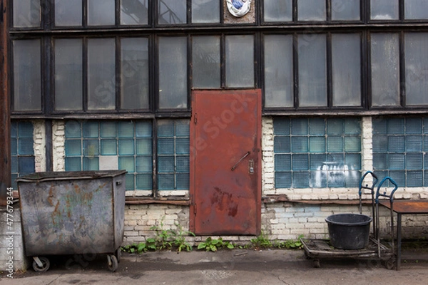Fototapeta old soviet constructivist industrial building exterior facade with red steel door, large windows, blue glass bricks. Rusty, trolly, weathered garbage container in front