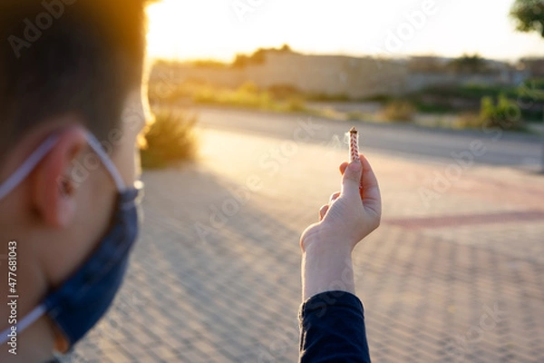 Fototapeta Boy holding a wick to play with firecrackers with facemask for covid protection