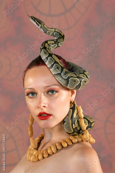 Fototapeta A beautiful woman poses nude for the camera. She is 
seen wearing a necklace and earrings made of peanuts. 
A python snake curls itself around her head. The model 
is seen nude in the photograph.
